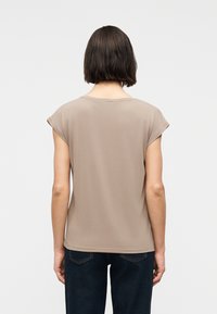 Γυναίκα με κοντά σκούρα μαλλιά, φορώντας μπεζ κοντομάνικη μπλούζα και σκούρο μπλε τζιν, που στέκεται με την πλάτη προς τα εμπρός σε απλό φόντο.