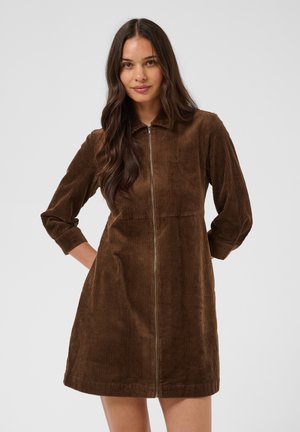 Femme aux longs cheveux foncés portant une robe en velours côtelé marron avec fermeture éclair à l'avant et manches trois quarts sur un fond clair uni.