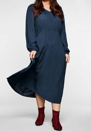 Nainen pitkähihainen laivastonsininen midimekko, jossa on kerätty vyötärö, pitelee helmaa, pukeutuneena viininpunaisiin nilkkureihin, yksinkertaisen taustan edessä.