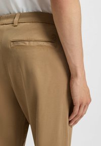 Bruna byxor med en slät textur, utrustade med en bakficka och subtilt logotypdetalj på midjebandet, visade från sidan.