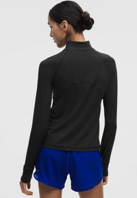 Pikad varrukad must sportlik topp kõrge kraega ja vormitud disainiga, koos siniste lühikeste pükstega. Sile tekstuur ja minimaalsed õmblusdetailid.