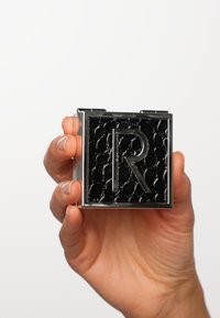 Schwarzer quadratischer Compact mit geprägtem R-Logo, glänzender Oberfläche und gepolsterter Textur. In einer Hand mit natürlichen Nägeln gehalten.