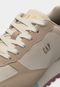 Gros plan sur une basket GAPS beige et crème avec des lacets blancs, des accents en daim et une semelle à motif.