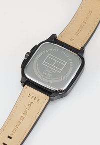 Montre carrée en acier inoxydable avec un fond gris, des accents noirs et un bracelet en cuir véritable avec des coutures visibles et du texte en relief.
