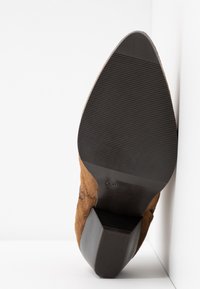 Bottine en daim marron avec un bout pointu, semelle noire texturée et talon carré, positionnée debout sur un fond blanc.