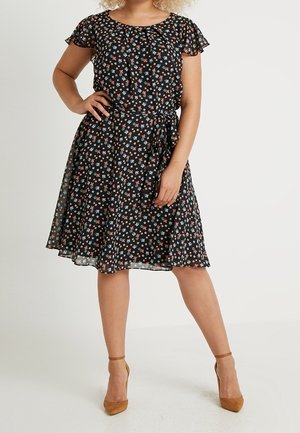 Kvinde iført en knælang sort kjole med små røde, hvide og blå blomsterprint, flagrende ærmer og brune ankelremssandaler.