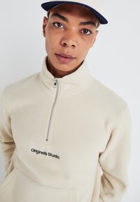 Beige fleece half-zip pullover with a front pocket and "Originals Studio" logo embroidered in black. Worn with a light cap.