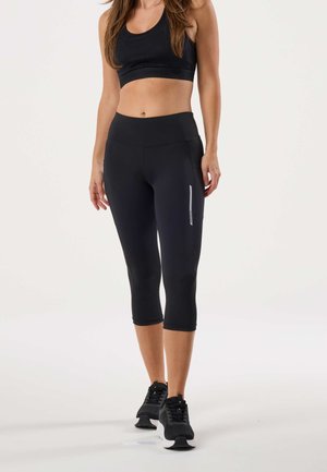 Vrouw gekleed in een zwarte sportbeha, zwarte cropped leggings met zak en reflecterende streep, en zwarte hardloopschoenen, staand tegen een effen achtergrond.