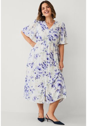 Kvinde smilende iført en hvid midi-kjole med lilla blomsterprint, korte udvidede ærmer og marineblå slingback-sko med lav hæl.