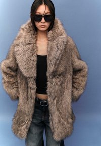 Cappotto oversize in pelliccia sintetica di color marrone chiaro con una trama morbida, ampio colletto e spalle scese. Indossato sopra un top nero senza maniche e jeans.