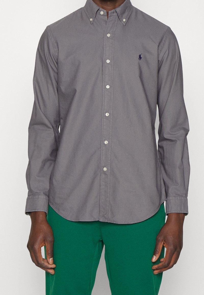 Szara koszula na guziki z długimi rękawami, z kołnierzykiem i małym logo na piersi. Tkanina ma gładką, lekko teksturowaną powierzchnię.