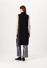 Μαύρο αμάνικο μακρύ γιλέκο με ψηλό γιακά, πλαϊνές σχισμές, συνδυασμένο με γυαλιστερά ασημί παντελόνια φαρδιάς γραμμής και μαύρα επίπεδα παπούτσια.