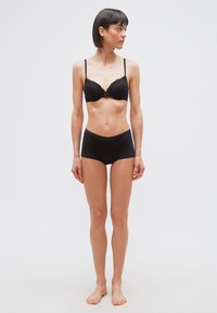 Γυναίκα με κοντά σκουρόχρωμα μαλλιά, στέκεται ξυπόλητη φορώντας μαύρο σουτιέν και μαύρο κοντόβρακο που ταιριάζει, κοιτώντας προς τα αριστερά της, πάνω σε απλό φωτεινό φόντο.