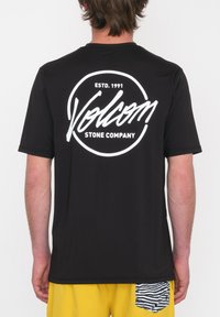 Camiseta negra de manga corta con un gran logo circular blanco en la parte posterior que incluye el texto "Volcom" y "ESTD. 1991", hecha de una tela suave.