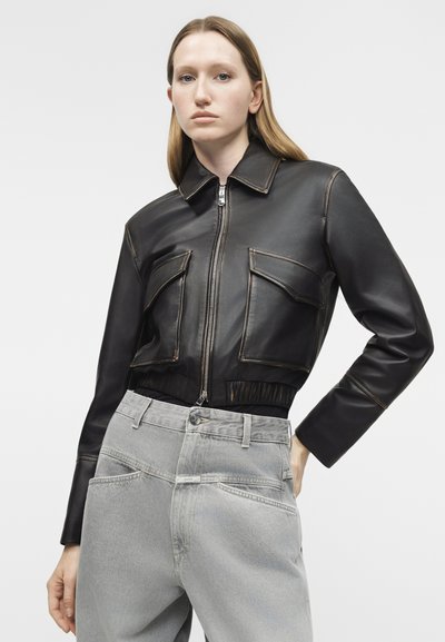 Woman wearing a black cropped leather jacket with front flap pockets and light gray high-waisted jeans, posing against a plain background.