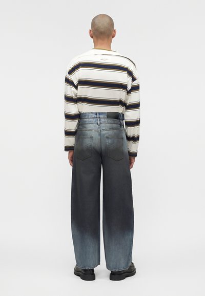 Άτομο με ξυρισμένο κεφάλι που φοράει λευκό ριγωτό πουκάμισο με μακριά μανίκια και δίχρωμα μπλε παντελόνια με φαρδιά πόδια, γυρισμένο με την πλάτη προς το γκρι φόντο.