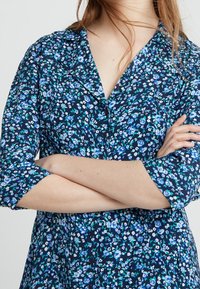 Sukienka koszulowa w kwiaty z granatowym tłem i niebieskimi oraz turkusowymi wzorami, z guzikami z przodu i rękawami 3/4.