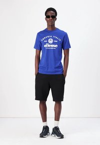 T-shirt azul de manga curta com texto branco, combinado com calções pretos. O modelo usa óculos de sol e sapatilhas pretas com meias brancas.