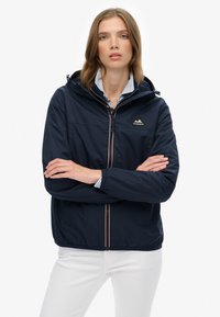 Femme aux cheveux châtain clair portant une veste à capuche bleu marine sur une chemise rayée, les bras croisés, se tenant devant un fond blanc.