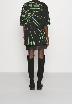 Osoba ubrana w czarno-zieloną koszulkę oversize z efektem tie-dye z napisem na górnej części pleców, czarną skórzaną spódnicę oraz czarne kozaki za kolano, stojąca tyłem.