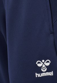 Mørkeblå stof med hvid hummel-logo, der indeholder en stiliseret bi og tekst, del af et tøjstykke med synlig lommesøm.