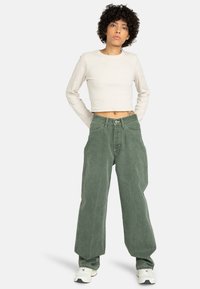 Haut long à manches longues beige clair coupé, associé à un jean en denim vert ample, avec une taille haute et une texture légèrement déchirée. Baskets blanches.
