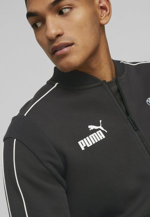 Jeune homme regardant à gauche, portant une veste zippée noire Puma avec des passepoils blancs et le logo BMW sur la poitrine.