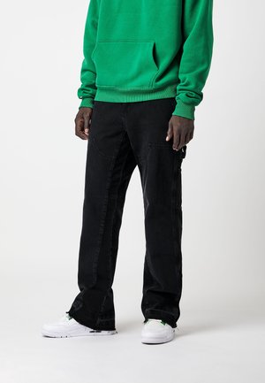 Personne portant un pantalon cargo noir, un sweat vert et des baskets blanches, debout devant un fond uni.
