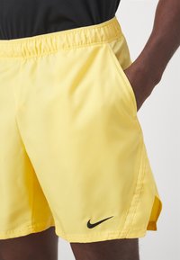 Keltaiset urheilushortsit, jotka on valmistettu sileästä kankaasta ja joissa on joustava vyötärönauha. Sivutaskut ja pieni musta Nike-logo vasemmassa helmassa.