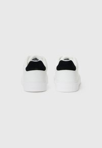 Adidași albi cu accente negre, având o parte superioară din piele netedă, guler cu umplutură și branding subtil pe călcâi. Talpă din cauciuc plat.
