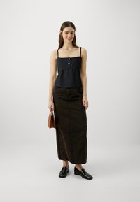 Svart crop top med justerbare stropper og tre knappfront, kombinert med et langt, mørkebrunt skjørt. Modellen holder en liten brun håndveske.