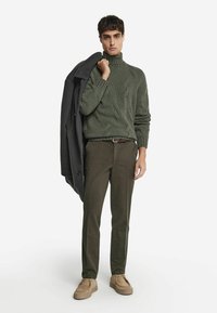 Grön stickad polotröja, mörkgrå rock över axeln, olivgröna byxor, beige slip-on skor med texturerad yta.