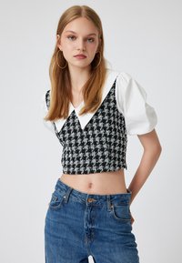Mustavalkoinen houndstooth-mallinen lyhyt toppi, jossa on valkoinen ylisuuri kaulus ja lyhyet hihat, yhdistettynä korkeavyötäröisiin sinisiin farkkuihin.
