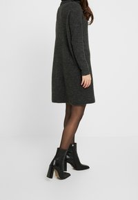 Vestito in maglia grigio carbone con una vestibilità comoda, texture a coste, maniche lunghe e un colletto alto, abbinato a stivaletti neri alla caviglia.