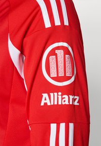 Sarkans sporta krekls ar baltām svītrām un logo. Teksts "Allianz" izdrukāts baltā krāsā. Materiāls ir teksturēts audums ar gaisa caurlaidīgu dizainu.