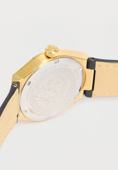 Dos de montre en or avec tête de Méduse gravée, bracelet en cuir noir et beige, et couronne en or sur fond blanc.