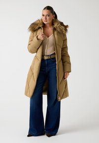 Cappotto imbottito beige con cappuccio foderato in pelliccia, dotato di zip frontale, tasche laterali e design a lunghezza ginocchio. Indossato sopra un maglione in maglia e jeans a gamba larga.