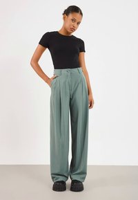 Kvinne står med hånden i lommen iført svart tettsittende kortermet topp, løse grønne høy midje bukser og svarte kraftige sko.