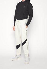 Svart hoodie med känguruficka, tillsammans med krämfärgade träningsbyxor med svarta diagonala ränder och elastisk midja. Vita sneakers.