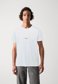 White cotton t-shirt featuring "GAP" in large cream letters and "Original" in red. Soft texture, classic crew neck design, short sleeves.