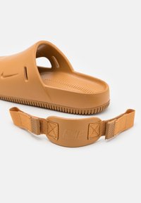 Nike sandaler i tan plast med åpen hæl og stroppfeste, med teksturert såle og logo på stroppen. Myk, glatt overflate.