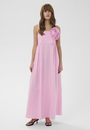 Mujer con vestido largo de color rosa sin mangas y una gran flor de tela en el hombro izquierdo, combinada con sandalias de tacón negras.