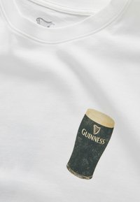 Maglietta bianca con un'immagine stampata di un bicchiere pint pieno di birra Guinness scura con una schiuma cremosa.