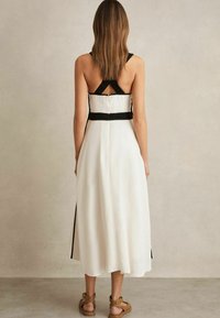 Weißes Midikleid mit schwarzen Trägern, ausgestattet mit einem kreuzgekreuzten Rückendesign und einem strukturierten Taillenbund. Glatte Stoffqualität, knöchellang, lässiger Stil.