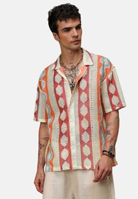 Koszula z krótkim rękawem, wzorzysta, wykonana z wielokolorowej siateczki. Charakteryzuje się geometricznymi wzorami w kolorze czerwonym, pomarańczowym, niebieskim i kremowym, w luźnym kroju.