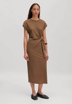 Frau steht, trägt ein braunes Midikleid mit seitlichem Knotendetail und schwarze flache Schuhe mit Fransen vor einem schlichten hellen Hintergrund.