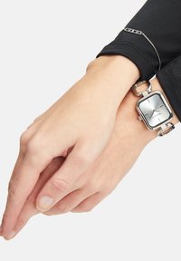 Silberne Metalluhr mit rechteckigem Zifferblatt, minimalen Stundenmarkierungen und einer Gliederarmband. Tragen auf einer Hand mit hellem Hautton.