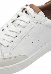 Zapatillas de cuero blancas con lados texturizados, cordones blancos planos y un acento en tono marrón en el talón. Presentan detalles perforados en la parte superior.