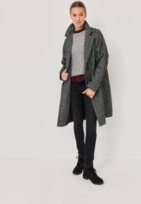 Grå texturerad kappa med hög krage, lager på lager över en grå topp och en vinröd skjorta, tillsammans med svarta skinny jeans och svarta snörboots.