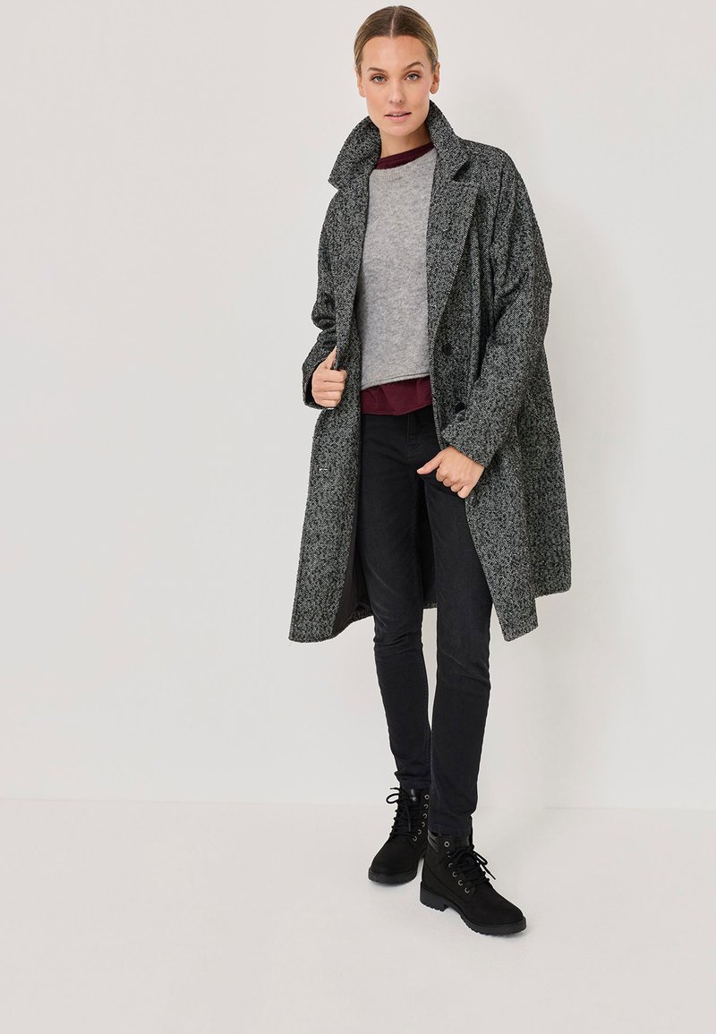 Grå texturerad kappa med hög krage, lager på lager över en grå topp och en vinröd skjorta, tillsammans med svarta skinny jeans och svarta snörboots.